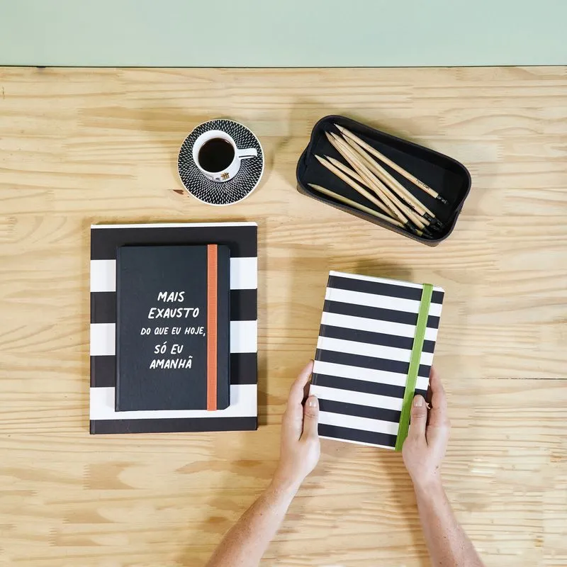 Uma pessoa com as mãos em uma agenda preto e branca, com uma xicara de café, outra agenda e uma caixa com lápis ao lado.