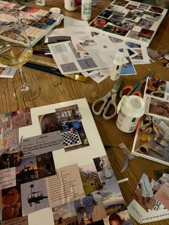 Foto de uma mesa cheia de recortes de revistas e papeis, cola tesoura e uma taça de vinho branco.